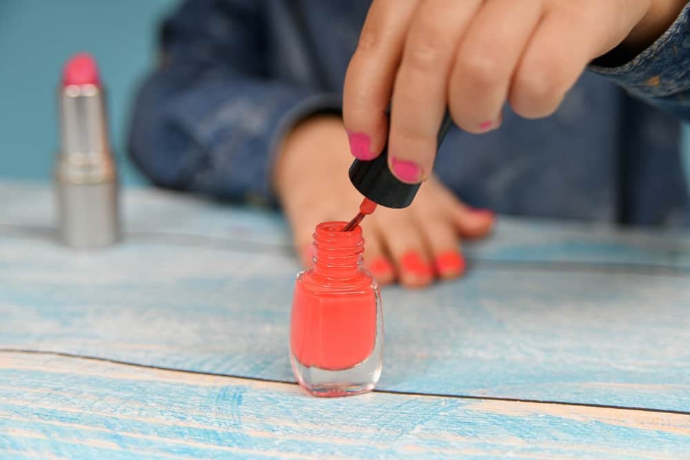 girl painted with her mother's makeup dips a brush into a bottle of nail polish